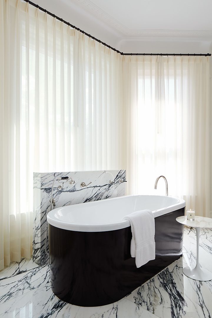 This is a luxurious primary bathroom featuring a freestanding black and white bathtub as the focal point. The bathroom is adorned with marble flooring and a marble backsplash behind the tub fixtures. Sheer white curtains filter natural light, creating a serene and spa-like atmosphere.