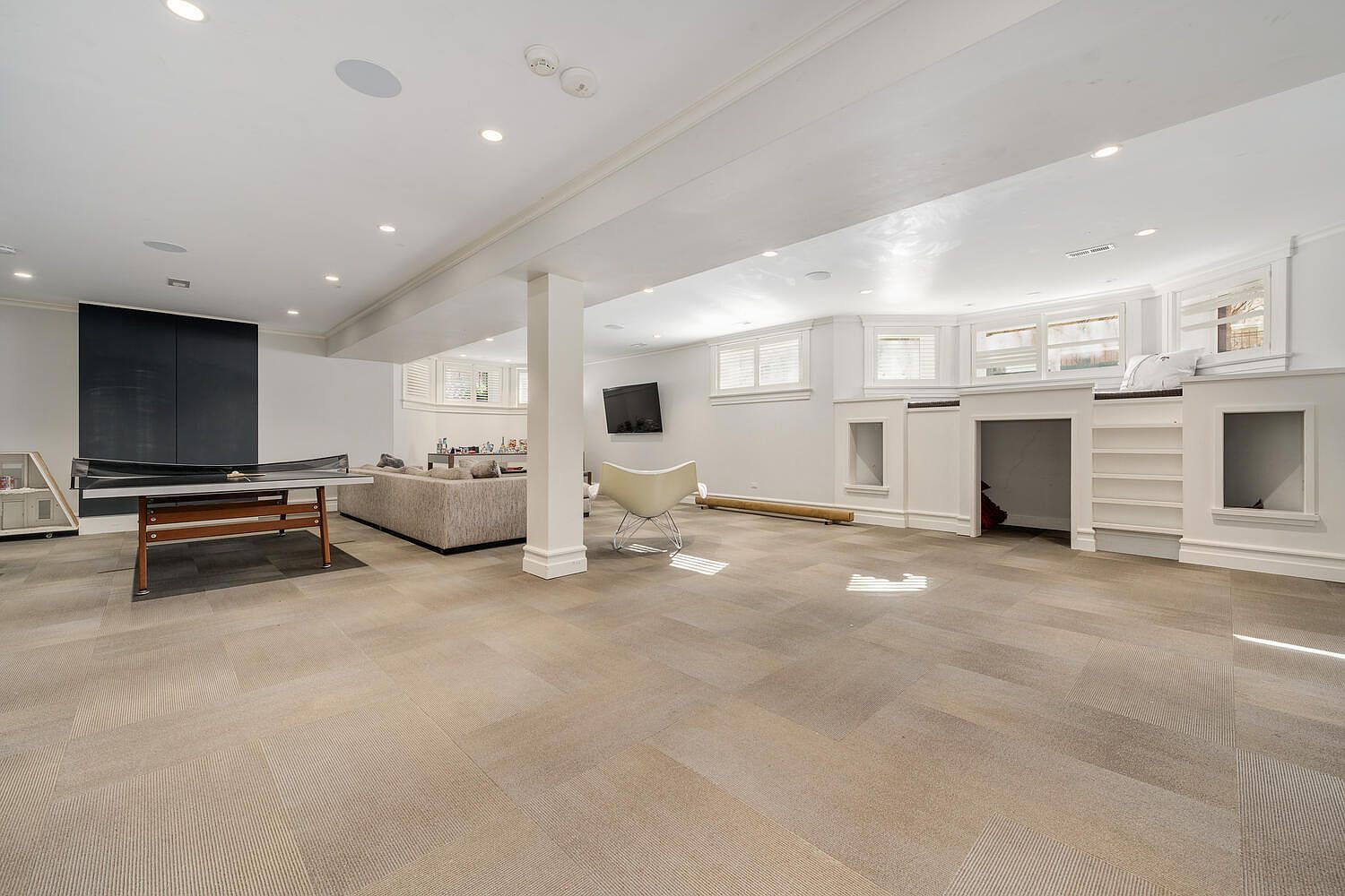 This is a spacious basement featuring a neutral-toned carpet, white walls, and recessed lighting. The area includes a ping pong table, a seating area with a sofa, and a built-in shelving unit with a faux fireplace. The room appears well-lit and offers ample space for recreation and relaxation.