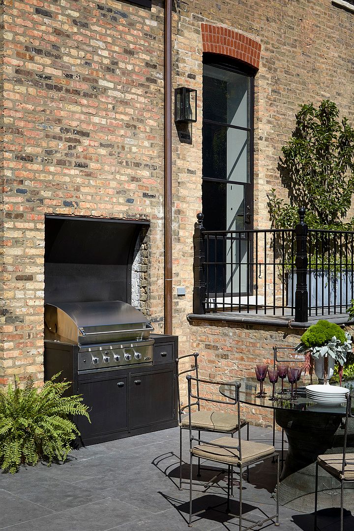 This image showcases an outdoor patio area featuring a built-in grill within a brick structure, complemented by a dining set with a glass table and wrought iron chairs. A balcony with a wrought iron railing is visible above, adding architectural interest to the brick facade. The scene suggests a sophisticated and inviting outdoor living space.