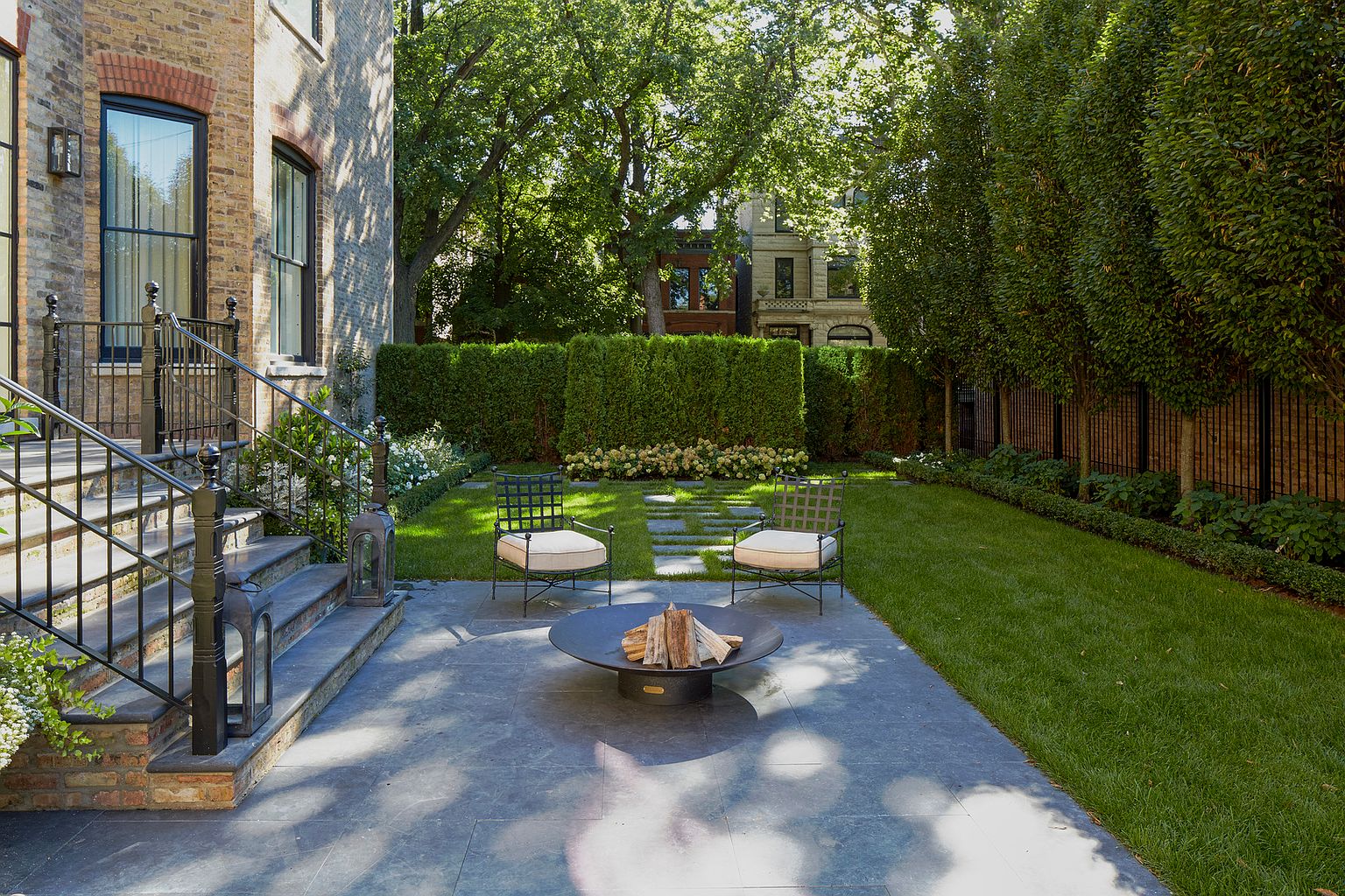 This is a beautifully landscaped backyard featuring a stone patio with a fire pit and two chairs, perfect for outdoor entertaining. The yard includes lush green grass, manicured hedges, and mature trees, creating a private and serene atmosphere. The architectural details of the house and the surrounding greenery enhance the property's appeal.