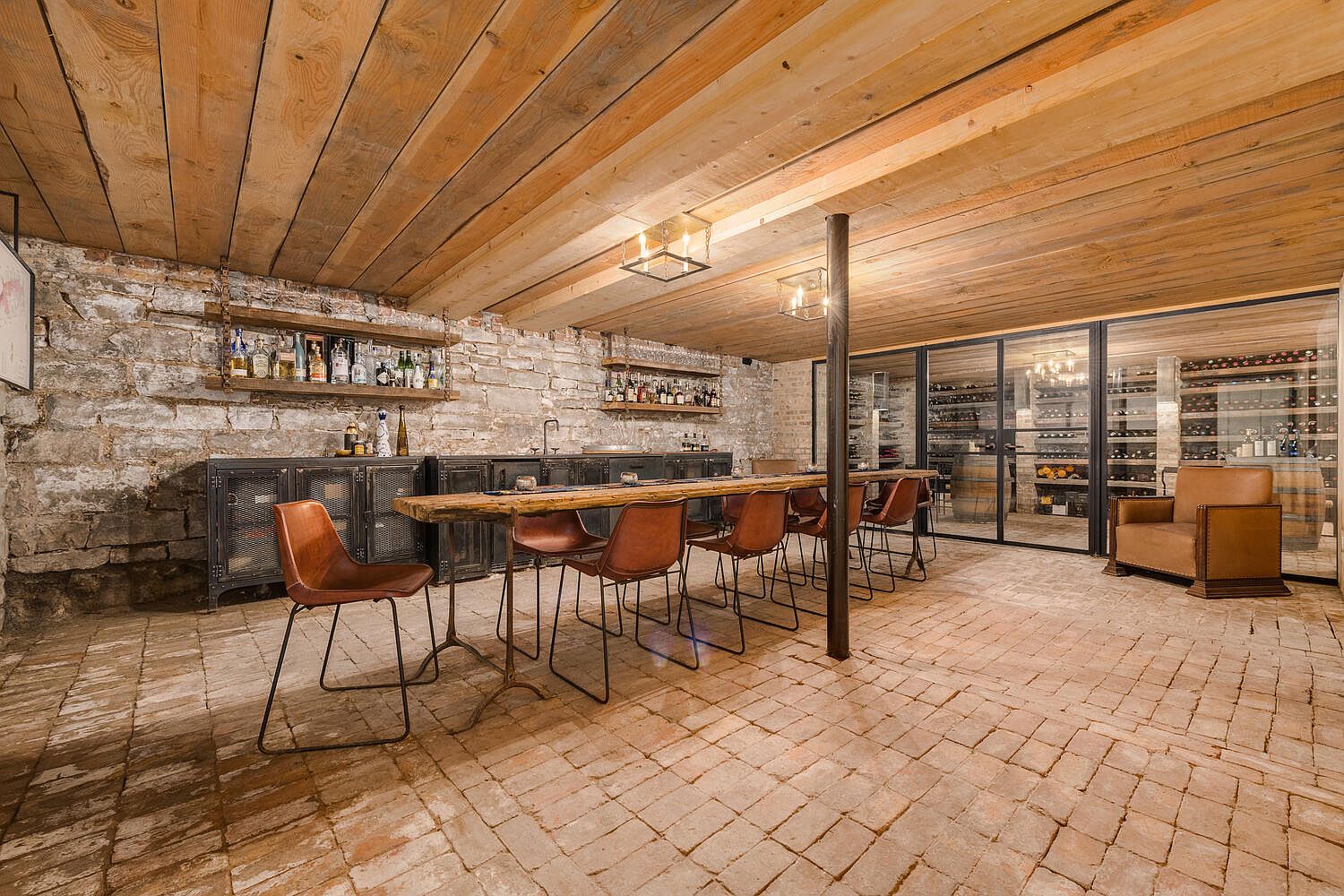 This is an interior shot of a wine cellar featuring a long wooden table with leather chairs, set against a stone wall with shelving displaying various bottles. A large wine storage unit with glass doors is visible in the background, and the room is lit by modern light fixtures, creating a rustic yet sophisticated ambiance. The floor is brick.