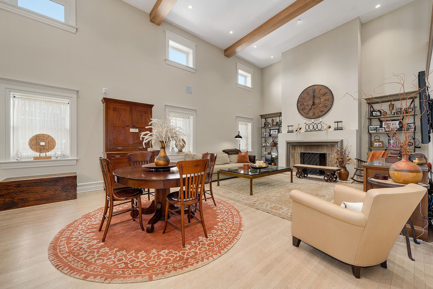 This is an interior shot of a living room featuring a high ceiling with wooden beams, a fireplace with a clock above it, and a seating area with a sofa and armchair. A round wooden dining table with chairs sits on an orange patterned rug, adding a touch of warmth to the space. The room is well-lit with natural light from the windows, creating a cozy and inviting atmosphere.
