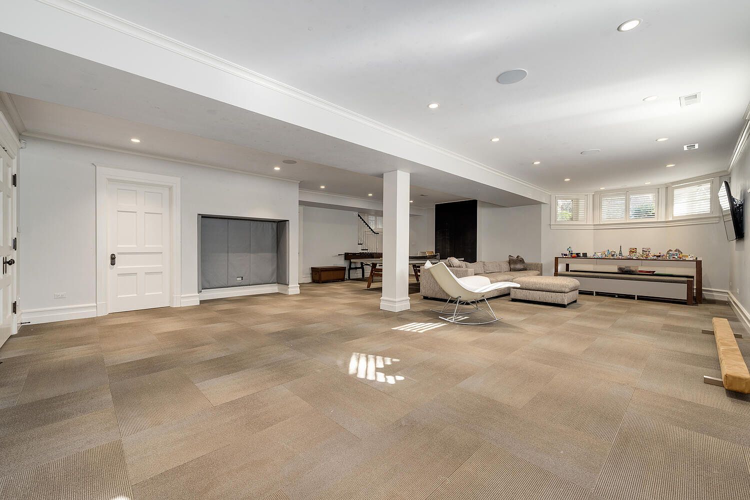 This is a spacious finished basement featuring neutral-toned carpet tiles and white walls. The area includes a seating area with a sectional sofa and a rocking chair, a console table with decorative items, and a pool table in the background. The room is well-lit with recessed lighting and natural light from the windows, creating a comfortable and inviting atmosphere.