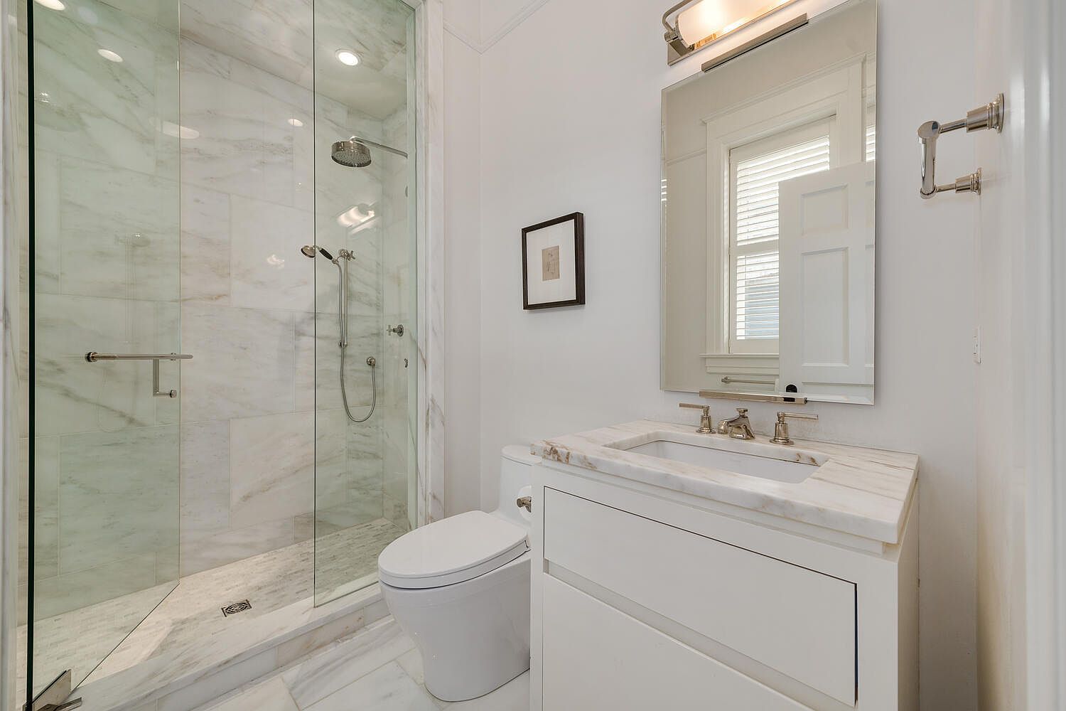 This is a well-lit bathroom featuring a glass-enclosed shower with marble tile, a white toilet, and a vanity with a marble countertop. The mirror reflects a window with blinds, and a framed picture hangs on the wall. The overall impression is clean, modern, and luxurious.