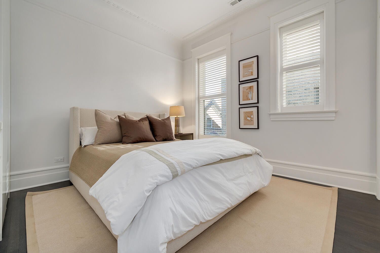 This is a bright and airy primary bedroom featuring a neutral color palette. The room is well-lit by natural light streaming through the windows, which are adorned with blinds. The bed is neatly made with a white duvet and several pillows, and a beige rug anchors the space, creating a cozy and inviting atmosphere.