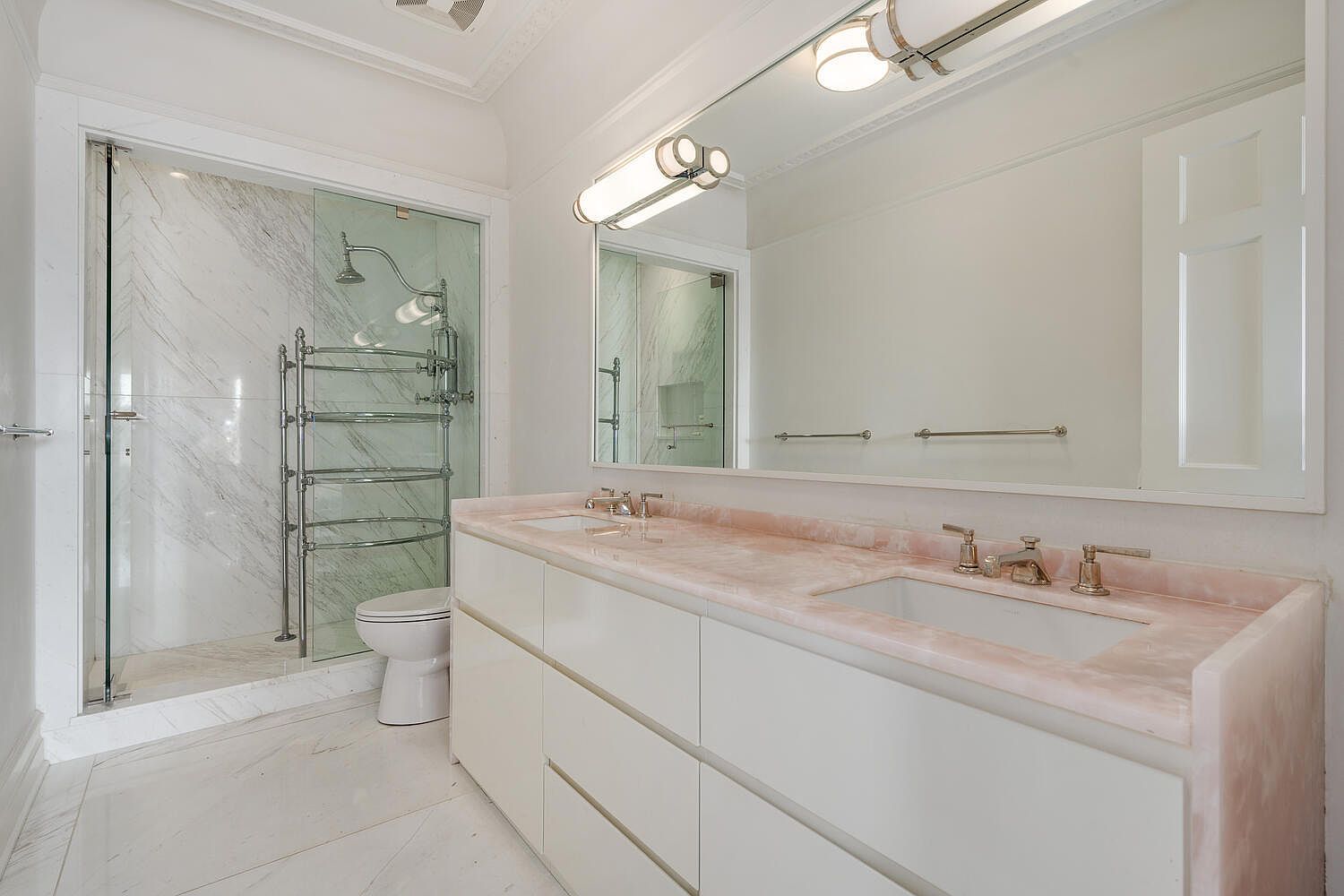 This is a well-lit primary bathroom featuring a double vanity with a pink marble countertop and white cabinets. A large mirror hangs above the vanity, reflecting the light fixtures and the shower area. The shower has glass doors and marble walls, and a heated towel rack is visible inside.