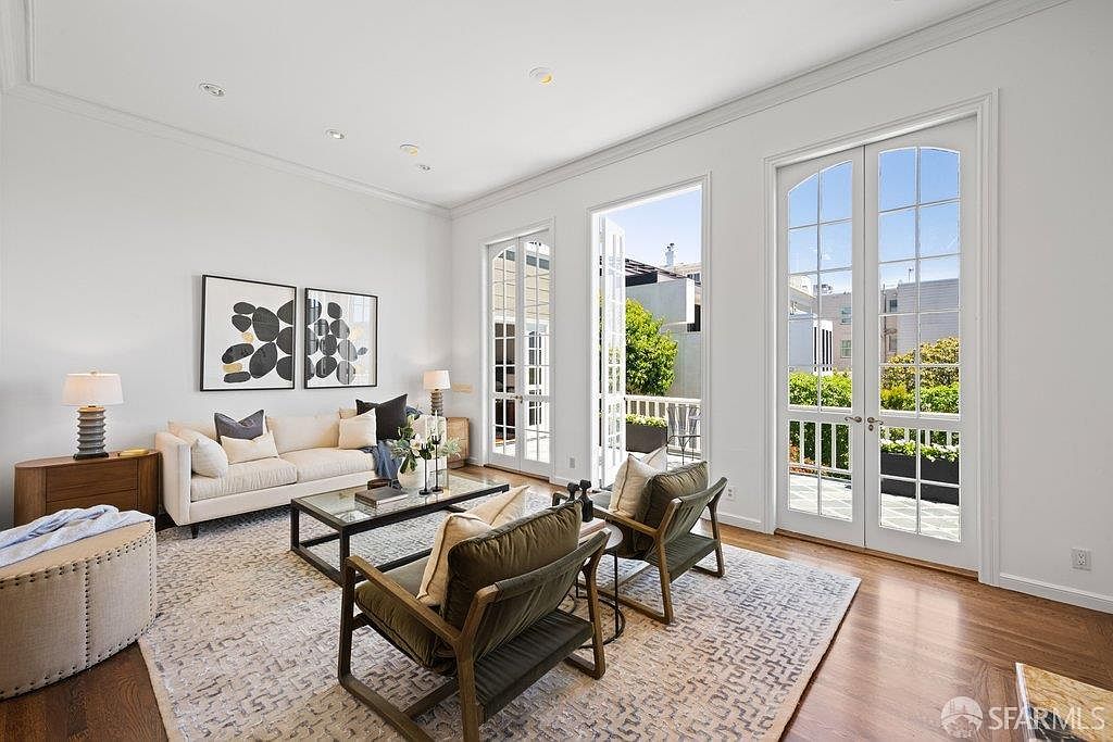 This bright and airy living room features high ceilings, hardwood floors, and a neutral color palette that creates an inviting atmosphere. The space is furnished with a cream-colored sofa, two olive-toned armchairs, and a glass-topped coffee table, all arranged on a large patterned area rug. Two sets of tall glass-paned French doors provide abundant natural light and offer a seamless transition to an outdoor balcony.
