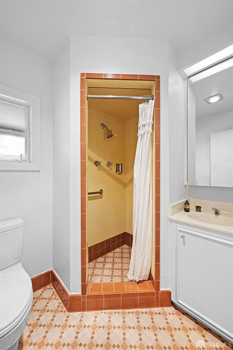 This bathroom features a compact layout with a shower stall enclosed by a white curtain, framed by distinctive orange-toned tile trim. The space includes a white vanity with a sink, a wall-mounted mirror, and a toilet, all set against a patterned tile floor. The overall aesthetic is functional and classic, with light-colored walls that brighten the small room.