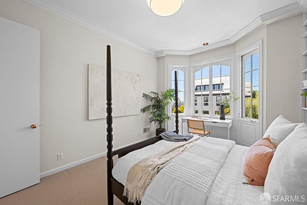 This bright and airy bedroom features a dark wood four-poster bed as the central focal point, complemented by neutral walls and light-toned carpeting. A bay window area provides a cozy nook for a desk and chair, allowing for ample natural light to fill the space. The room is accented by a large piece of abstract art and a potted plant, creating a serene and inviting atmosphere.