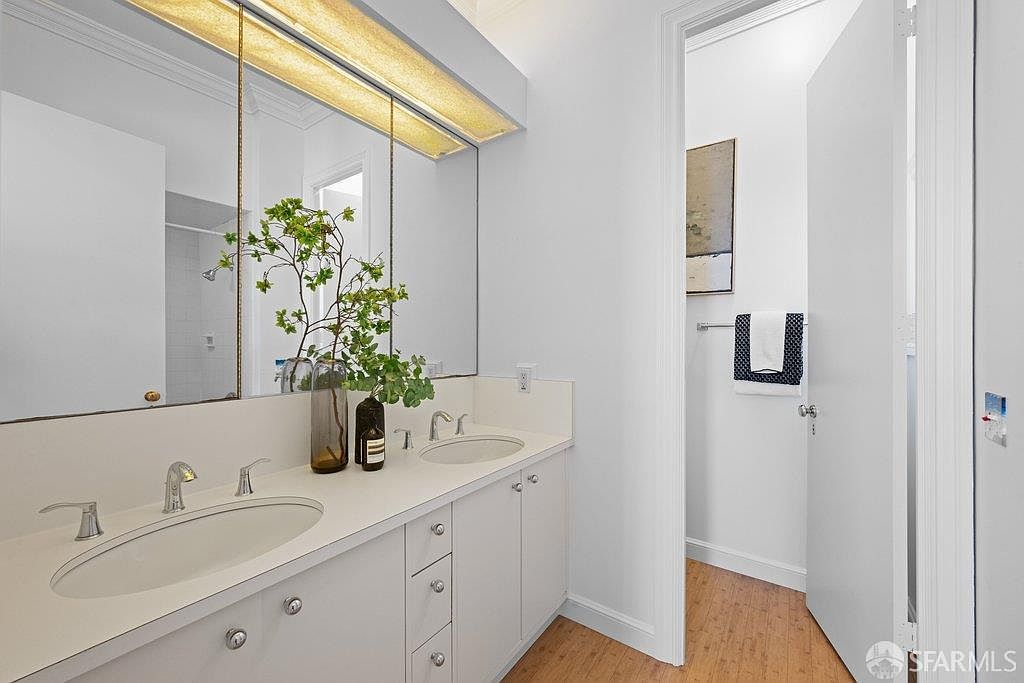 This bright and clean bathroom features a dual-sink vanity with white cabinetry and a large, full-width mirror that enhances the sense of space. The room includes a separate water closet area visible through an open doorway, and the light-toned wood flooring adds a warm, natural touch. A decorative plant arrangement on the counter provides a fresh, inviting accent to the minimalist design.