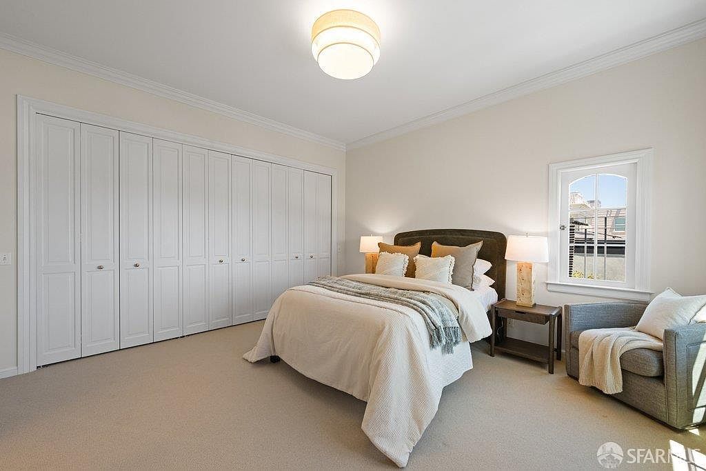 This bright and neutral bedroom features a queen-sized bed centered against a dark headboard, flanked by two matching nightstands with table lamps. A large wall of white, multi-panel closet doors provides ample storage, while a cozy armchair sits in the corner near a window that allows natural light to fill the room. The space is finished with light-toned carpeting and a simple, modern ceiling light fixture, creating a clean and inviting atmosphere.