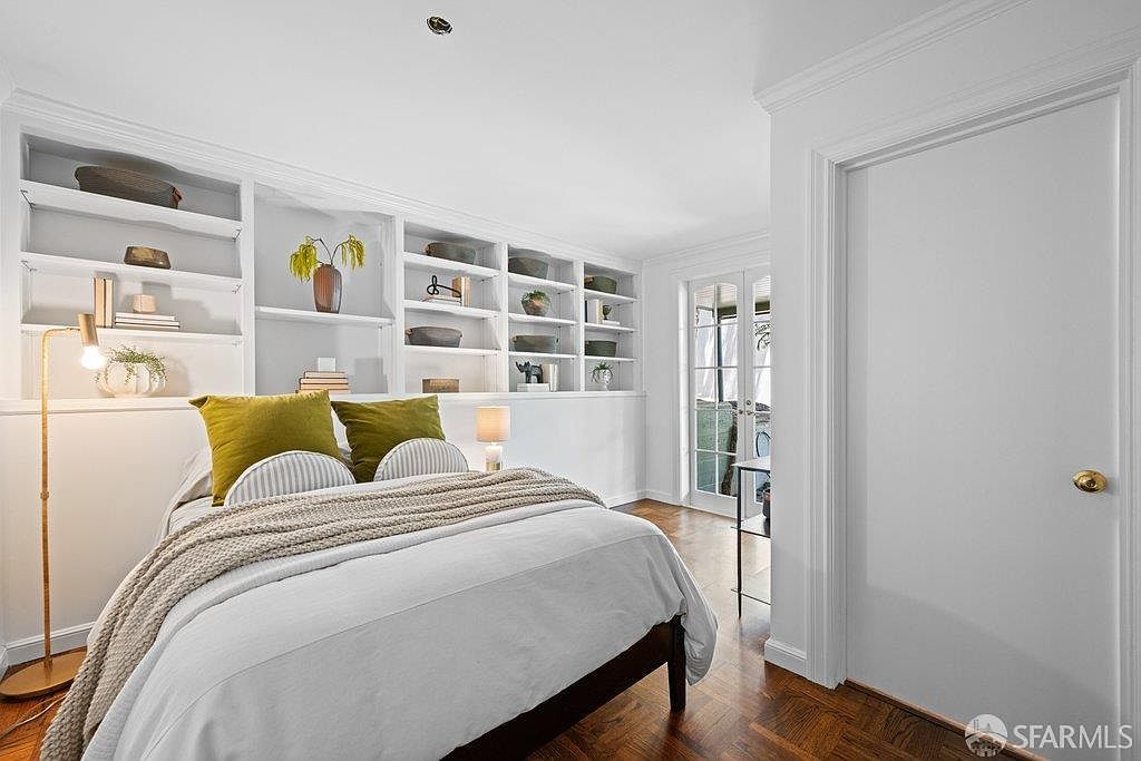 This bright and inviting bedroom features a queen-sized bed with crisp white bedding and olive green accent pillows. A large, built-in white shelving unit spans the wall behind the bed, providing ample display space for decor and books. The room is finished with warm-toned wood flooring and includes a set of glass-paned doors that lead to an outdoor area, allowing for plenty of natural light.