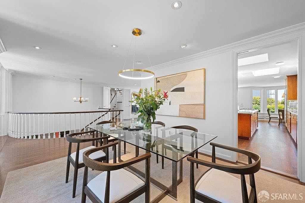 This bright and airy dining room features a contemporary glass-topped table surrounded by six modern chairs with dark wood frames and light-colored upholstery. A large, abstract piece of art hangs on the wall above the table, while a minimalist circular pendant light provides a focal point overhead. The room opens into a kitchen area and overlooks a staircase with a white railing, creating an open-concept feel with hardwood flooring throughout.