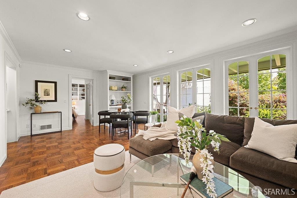 This bright and airy living room features rich, patterned wood flooring and a neutral color palette that creates a welcoming atmosphere. A large, comfortable sectional sofa faces a glass-topped coffee table, while a dining area with a built-in shelving unit is visible in the background. Multiple sets of French doors allow for abundant natural light and provide a seamless connection to the outdoor space.