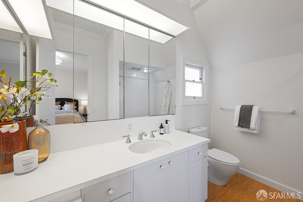 This bright and clean bathroom features a white vanity with a single sink, topped with a light-colored counter and polished metal fixtures. A large, multi-panel mirrored medicine cabinet spans the wall above the vanity, reflecting the adjacent bedroom, while a white toilet sits beside a towel rack holding folded linens. The room is finished with light-toned wood flooring and white walls, creating a simple and functional aesthetic.