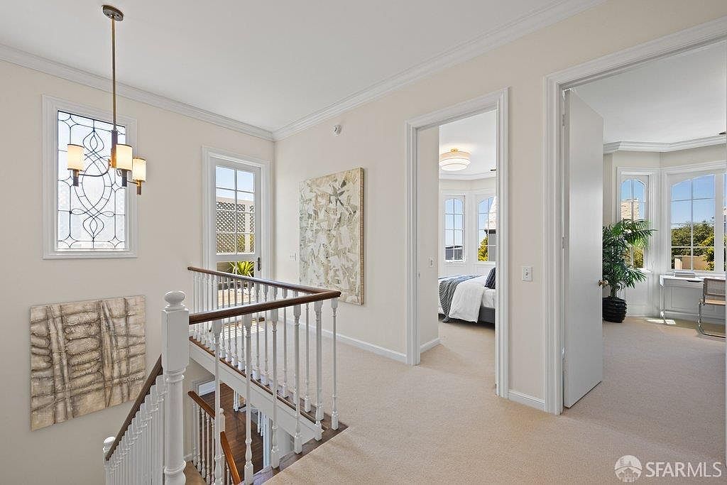 This bright, neutral-toned landing area features a staircase with a white railing and a wooden handrail, leading to an upper level. The space is decorated with large abstract art pieces and illuminated by a decorative chandelier, with doorways opening into a bedroom and a sunlit office space. The perspective is from the top of the stairs, looking across the landing toward the adjacent rooms.