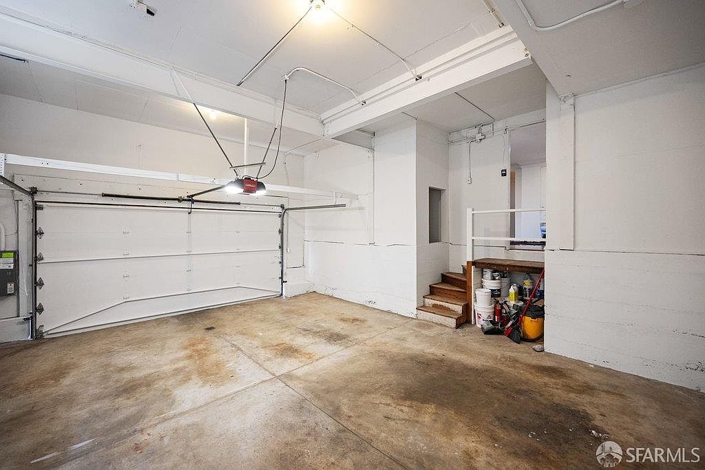 This image shows a spacious, utilitarian garage with a white sectional door and exposed ceiling infrastructure. A set of wooden stairs leads from the concrete floor up to a doorway, with a small storage area tucked underneath the landing. The space is brightly lit by overhead lighting, highlighting the concrete floor and painted block walls.
