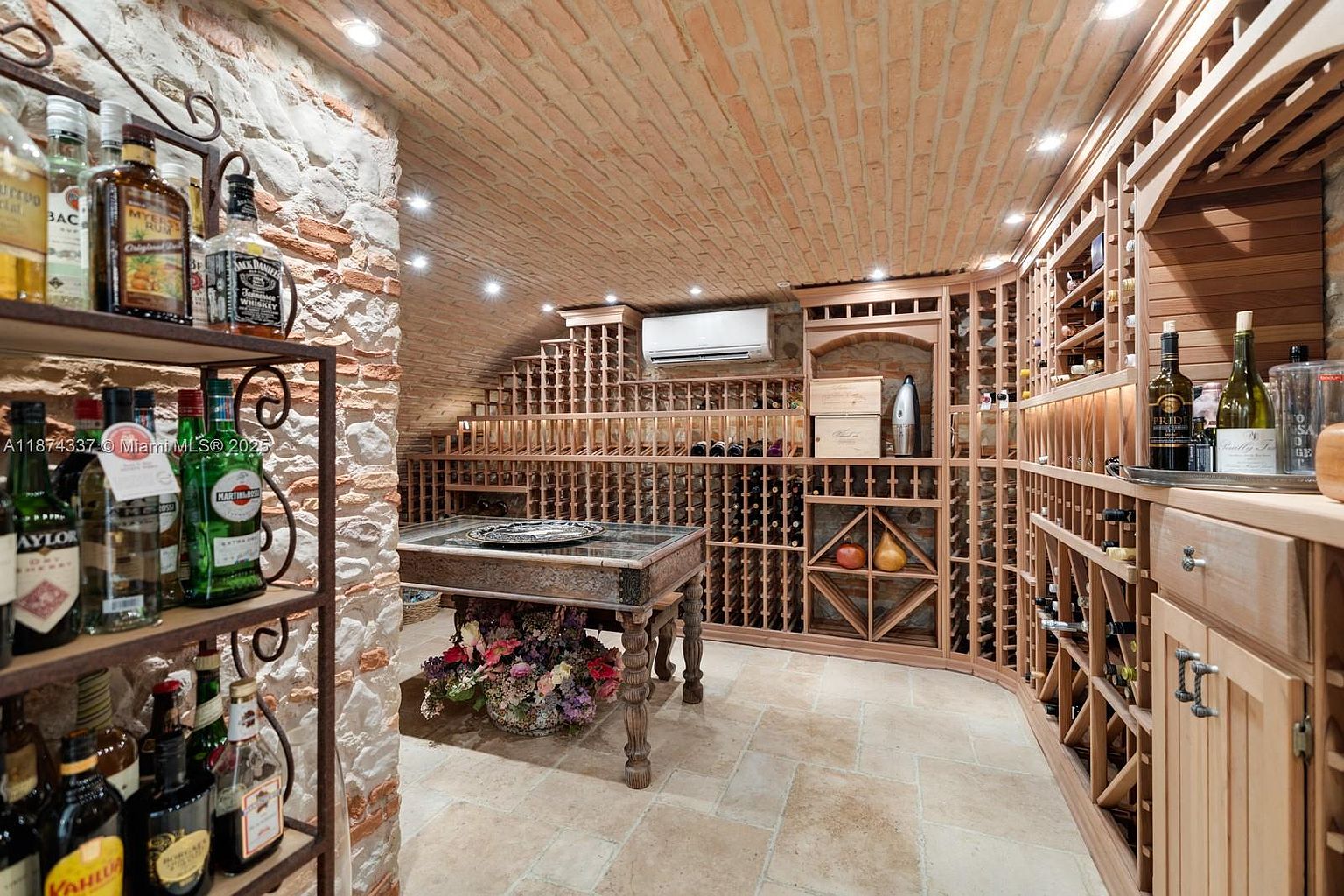 This is a well-appointed wine cellar featuring brick walls and ceiling, custom wooden wine racks, and a central table with floral arrangements. The room is illuminated by recessed lighting, creating a warm and inviting atmosphere. The design emphasizes both functionality and aesthetic appeal, making it a standout feature for any wine enthusiast.