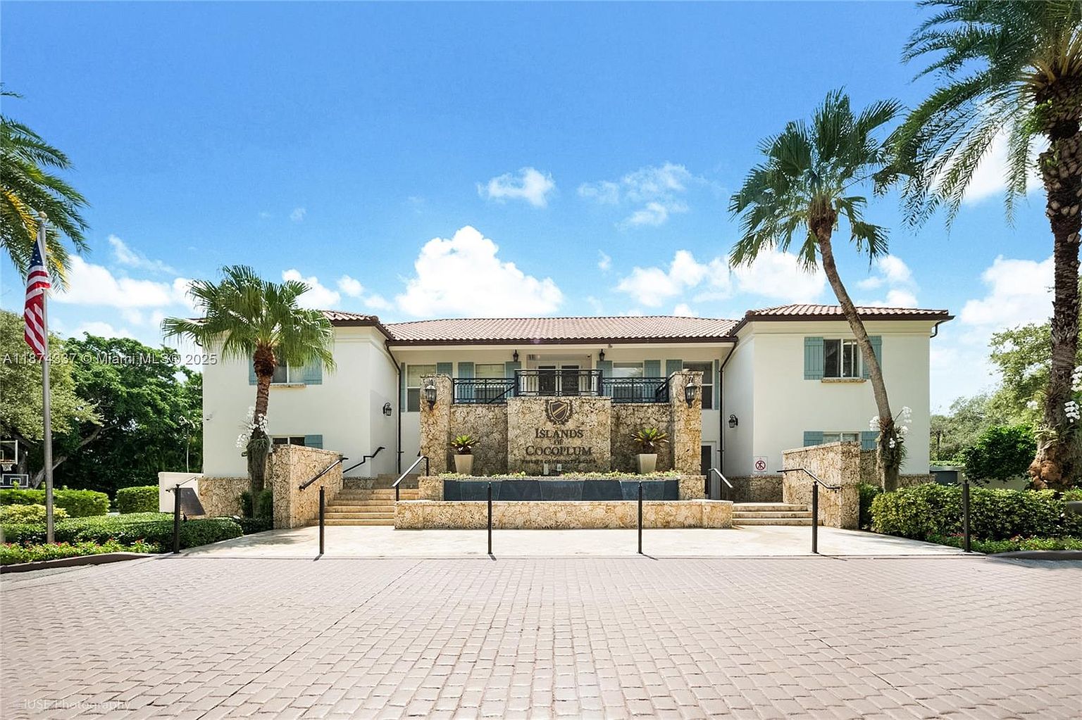 The image showcases the front exterior of a building complex, possibly a condominium or residential community, under a bright sky with scattered clouds. The building features a symmetrical design with a stone facade at the entrance, flanked by palm trees and well-maintained landscaping. A paved driveway leads up to the entrance, creating a welcoming and upscale impression.