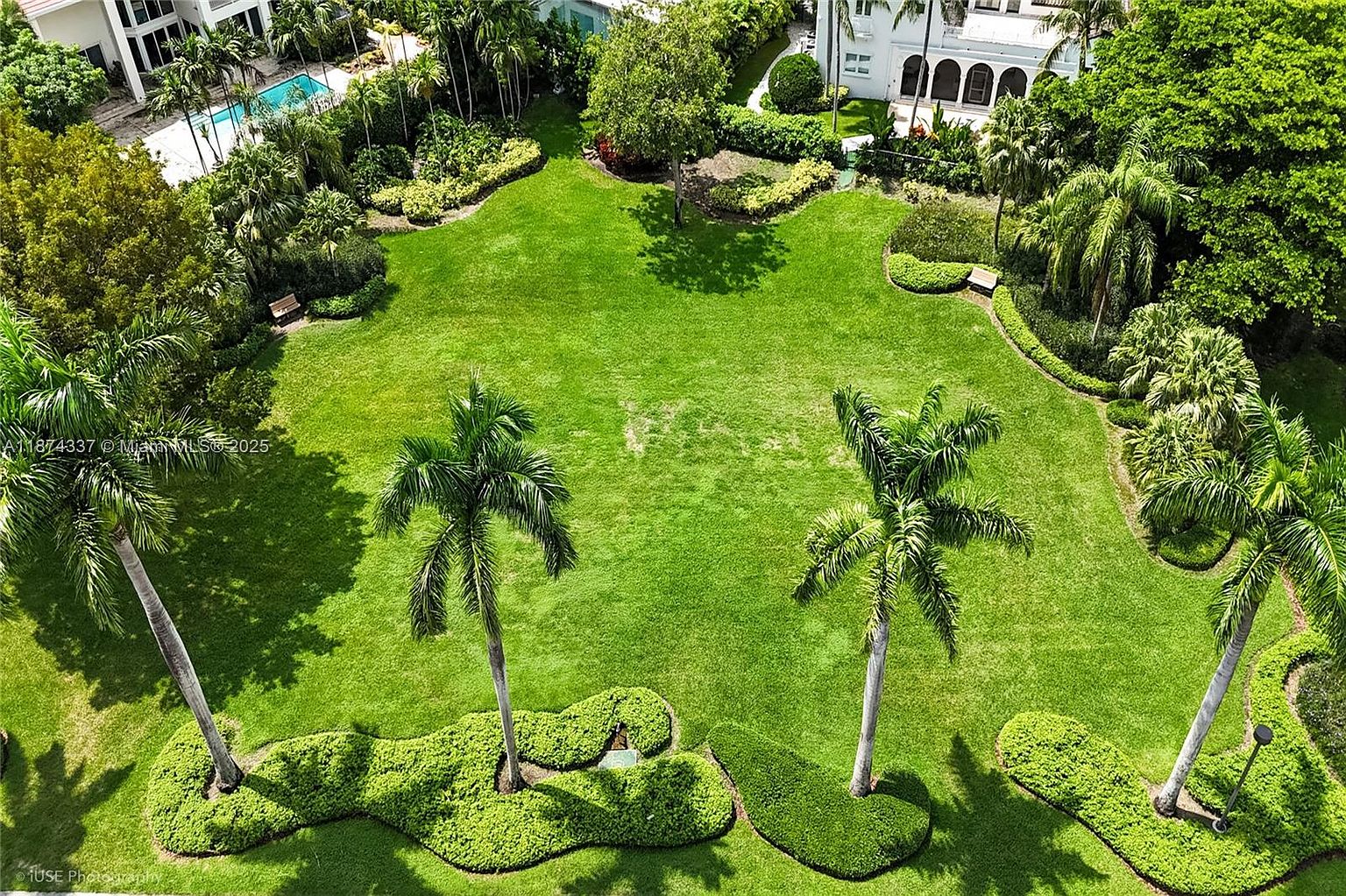 This is an aerial view of a meticulously maintained yard or garden. The lush green lawn is punctuated by mature palm trees and carefully manicured shrubbery. The property also features a swimming pool and a large house, suggesting a high-end residential estate.