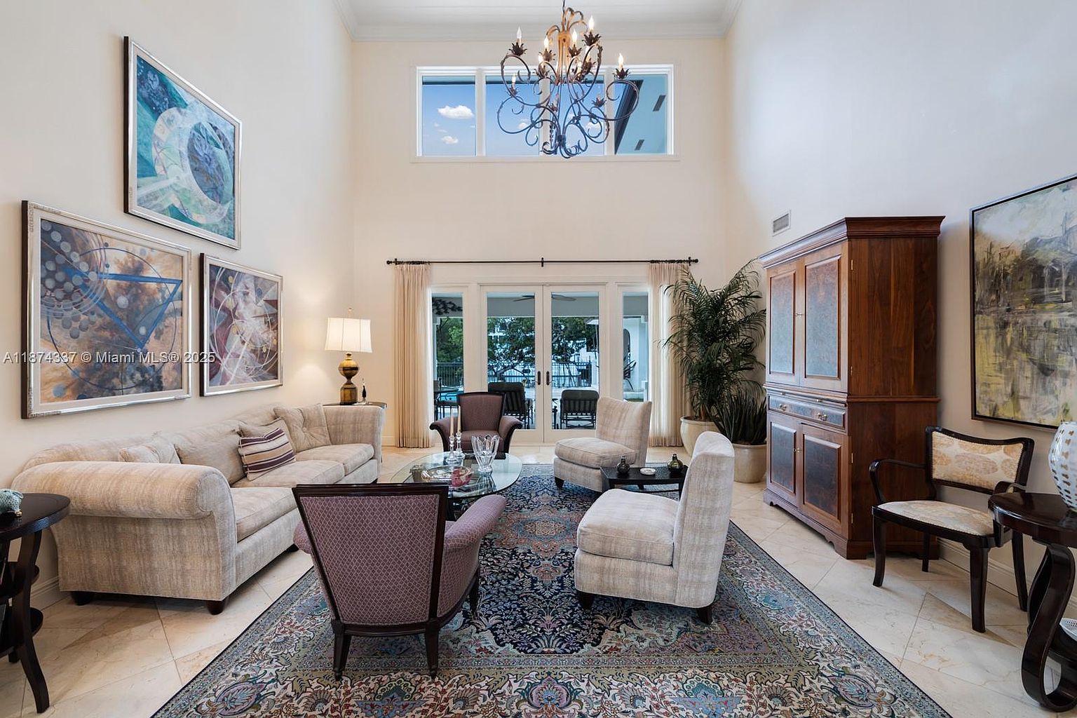 This is an interior shot of a living room featuring a high ceiling with a chandelier, a large area rug, and several pieces of furniture including a sofa, armchairs, and a wooden cabinet. Artwork adorns the walls, and natural light streams in through the windows and glass doors, creating a bright and inviting atmosphere. The room exudes a sense of elegance and comfort.