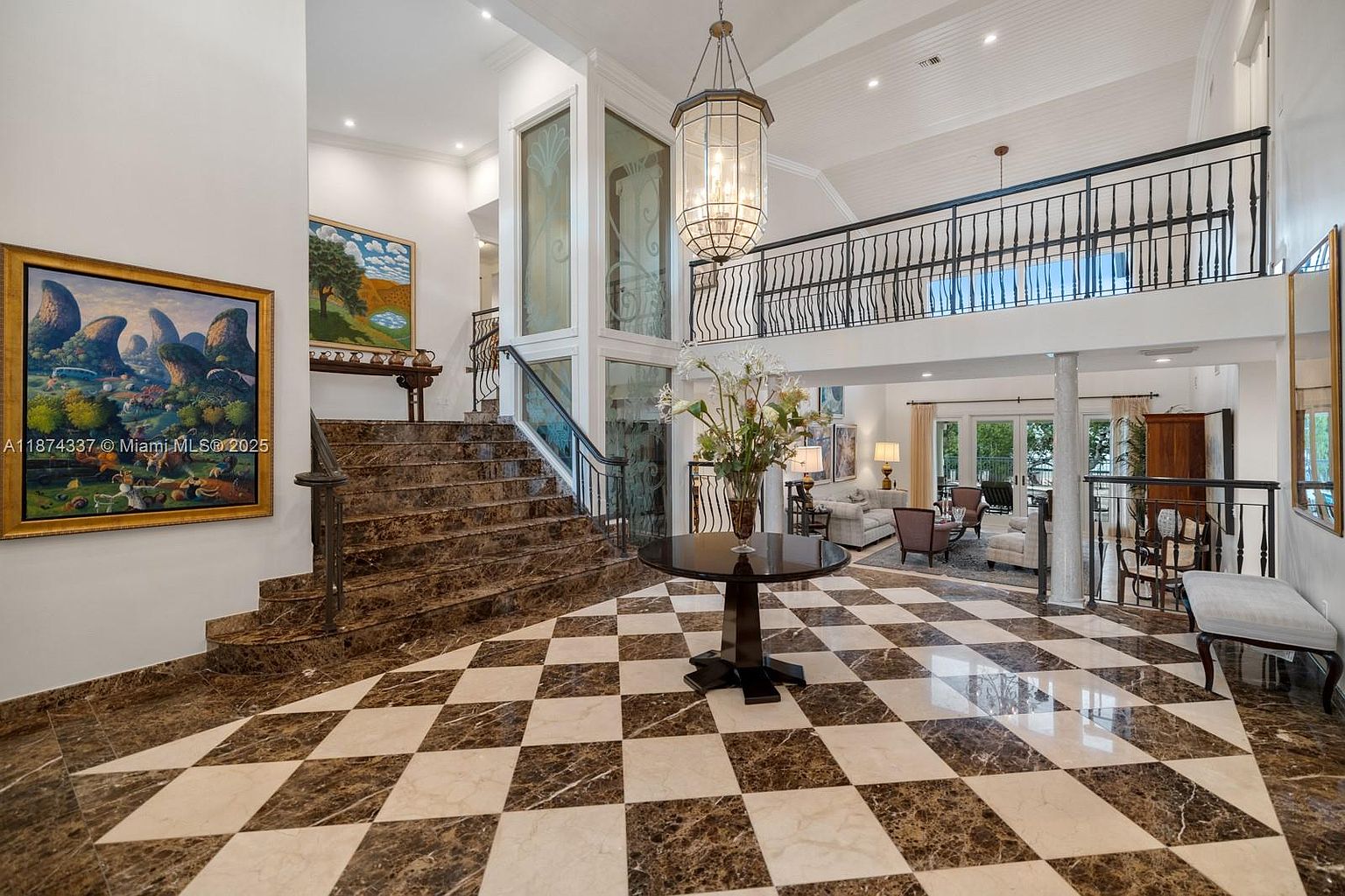 This grand foyer features a striking checkered marble floor, a sweeping staircase with marble steps and wrought iron railings, and a large, ornate chandelier. A second-floor balcony overlooks the entrance, adding to the sense of spaciousness and luxury. Artwork adorns the walls, and a floral arrangement sits on a round table in the center of the room, creating a welcoming and elegant first impression.