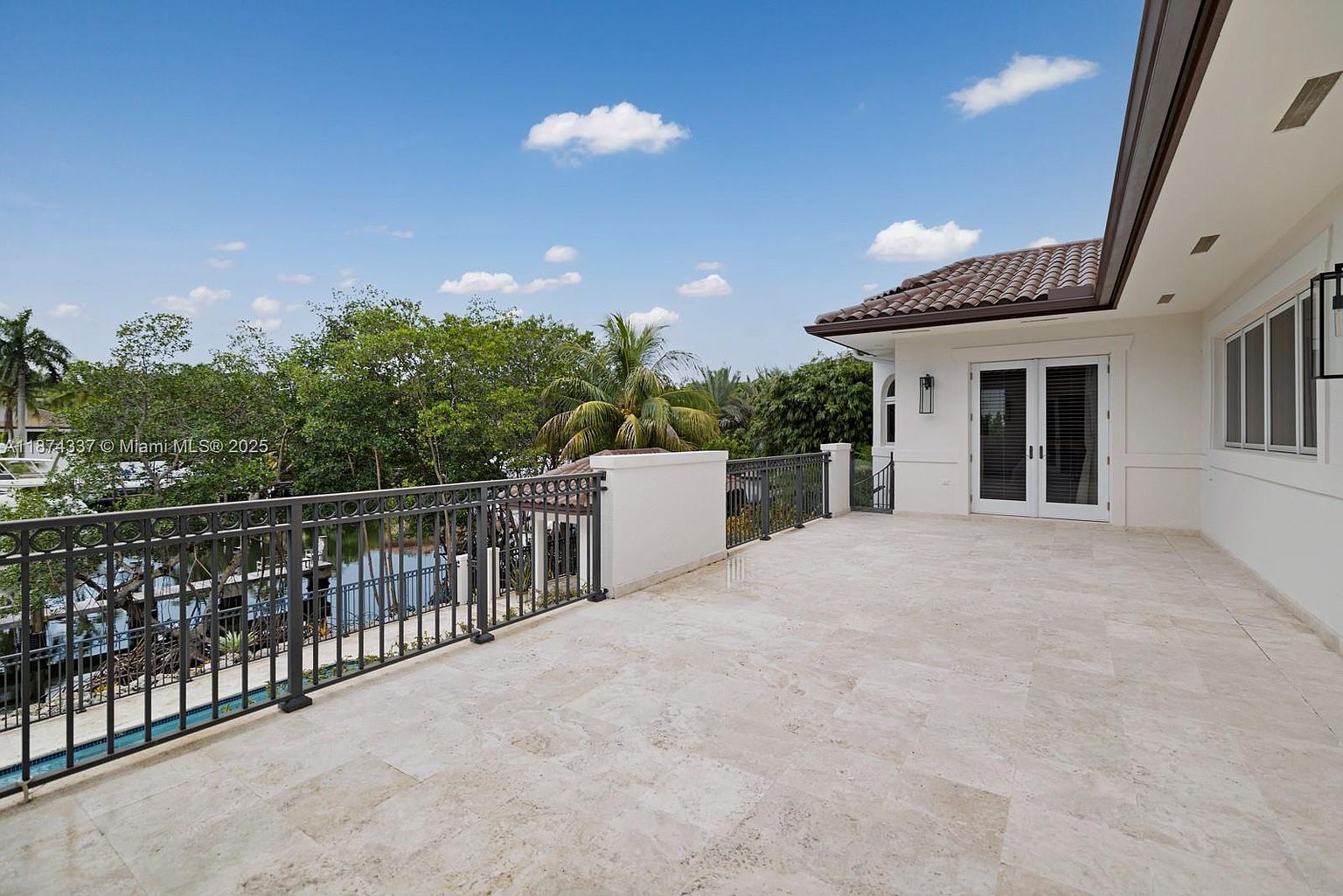 This image showcases a spacious balcony with a light-colored stone tile floor and a decorative metal railing. The balcony offers a view of lush greenery and a glimpse of a pool below. The building's exterior is painted white, and the balcony features double doors leading inside, suggesting a seamless indoor-outdoor living experience.