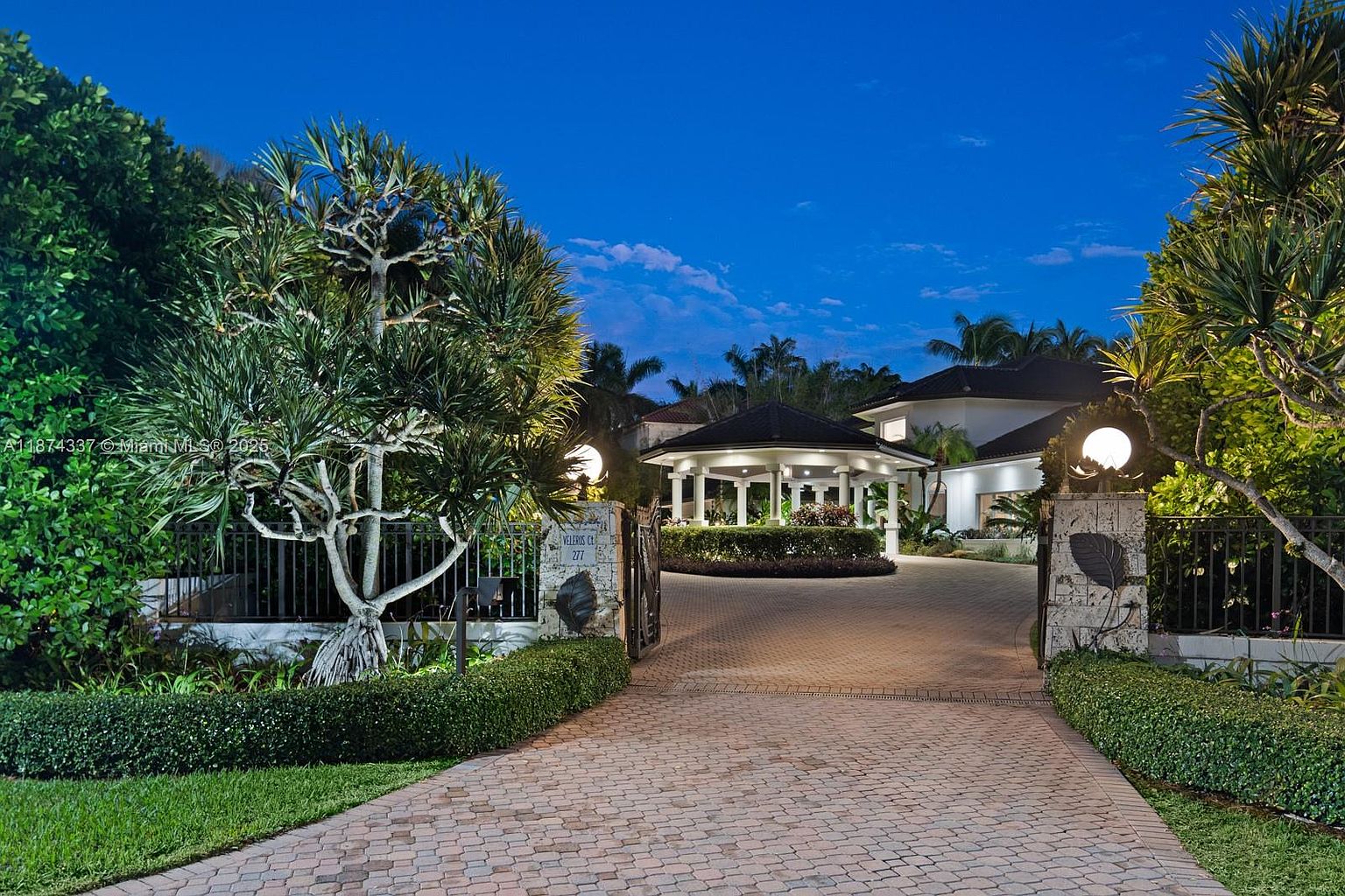 The image showcases a grand entryway to a property, featuring a brick-paved driveway leading to a circular structure with white columns and a dark roof. The entrance is flanked by stone pillars with decorative lighting and lush greenery, creating an inviting and luxurious first impression. A wrought-iron gate adds a touch of security and elegance.