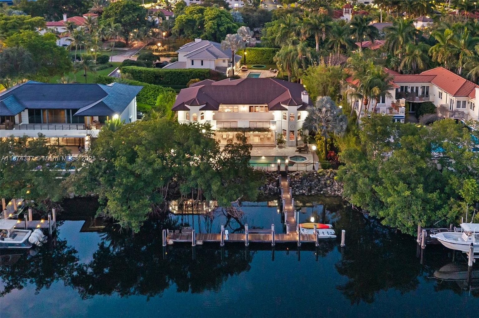 This aerial view showcases a luxurious waterfront property featuring a large two-story house with a red tile roof, a private dock extending into the water, and lush landscaping. The property includes a pool and spa area, enhancing its appeal as a high-end residence. The surrounding neighborhood is filled with mature trees and other upscale homes, creating a sense of exclusivity and tranquility.
