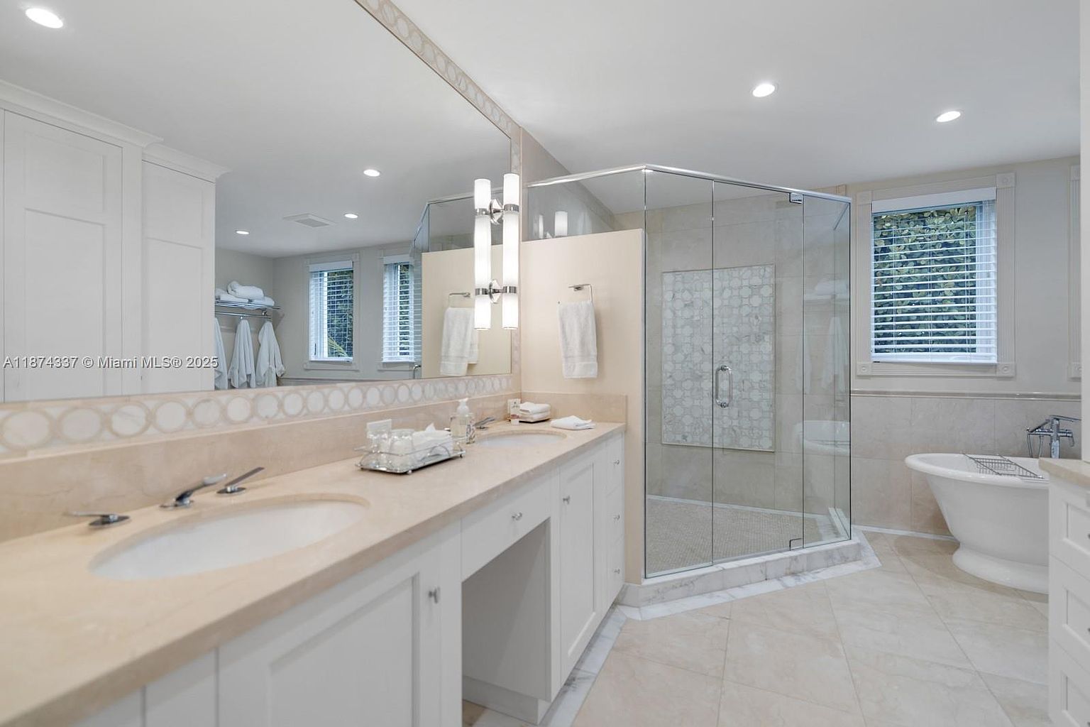 This is a well-lit primary bathroom featuring a double vanity with a light beige countertop and white cabinetry. A large mirror spans the length of the vanity, reflecting natural light from the windows. A glass-enclosed shower and a freestanding bathtub are also visible, contributing to the bathroom's luxurious and spa-like atmosphere.