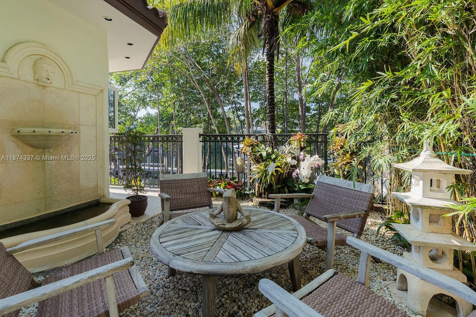This image showcases a tranquil garden patio area, featuring a round wooden table surrounded by comfortable wicker chairs. A serene water feature with a Buddha head is visible in the background, complemented by lush greenery, including bamboo and various plants. The setting evokes a sense of peace and relaxation, making it an ideal outdoor space for a home.