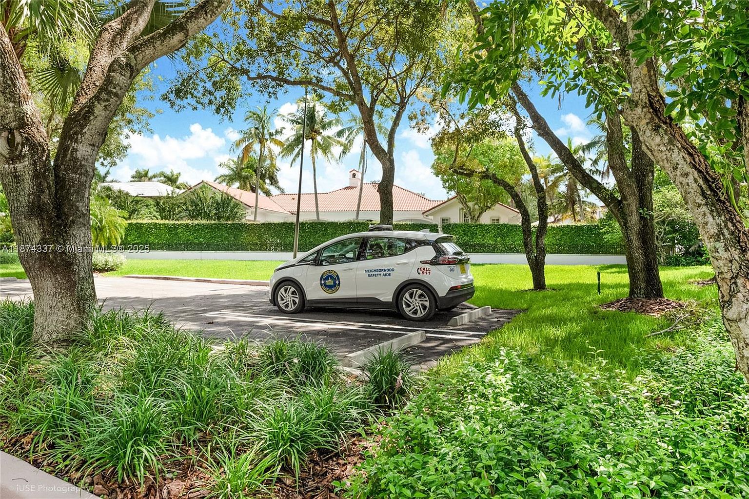 The image showcases the side view of a white neighborhood safety aide vehicle parked in a designated spot, framed by lush greenery and mature trees. In the background, a well-maintained house with a red tile roof is visible behind a tall, manicured hedge, creating a sense of privacy and security. The scene is bathed in natural light, emphasizing the peaceful and secure environment of the neighborhood.