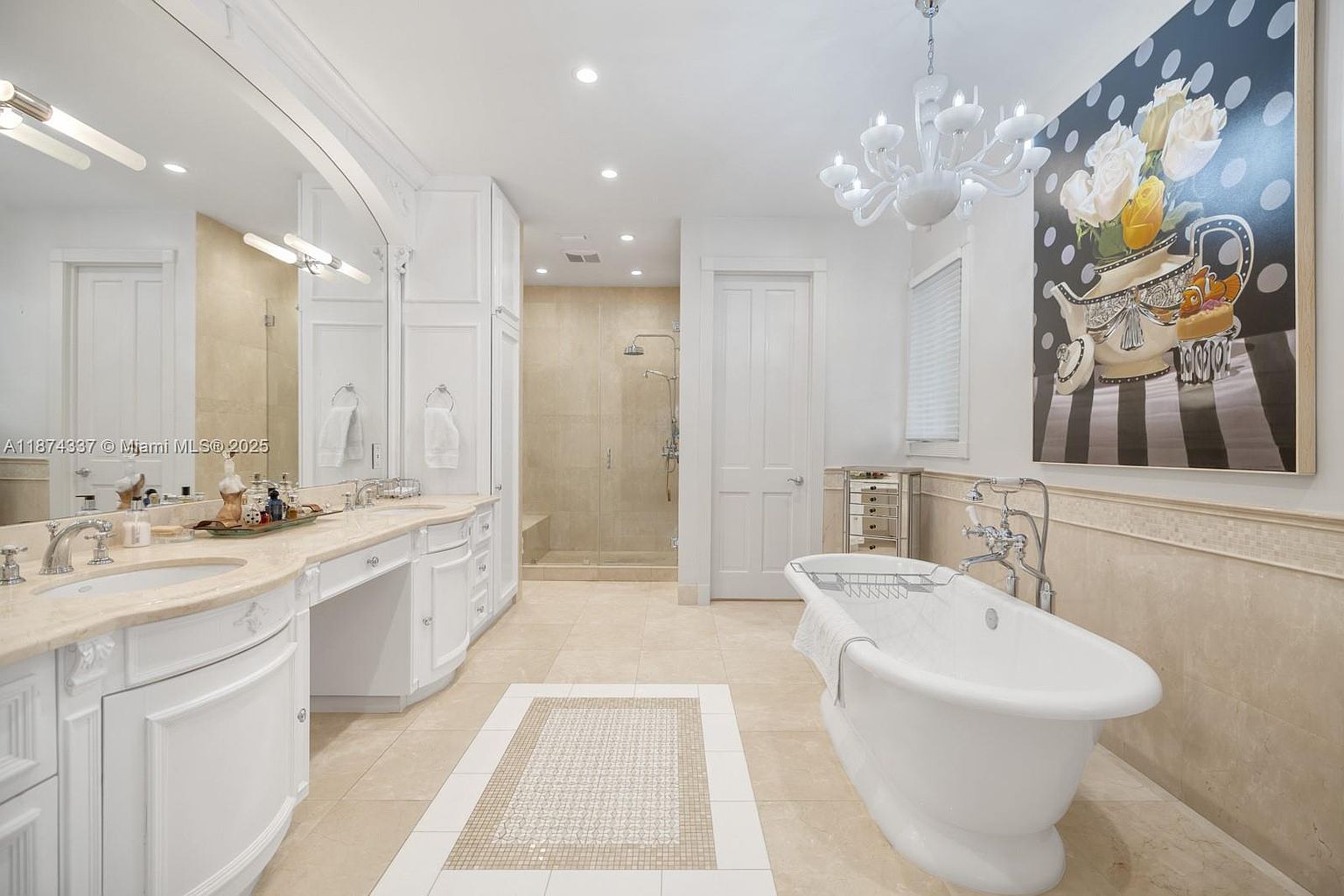 This is a luxurious primary bathroom featuring a freestanding bathtub, a double vanity with marble countertops, and a separate shower area. The room is decorated in a neutral color palette with white cabinetry and beige tile flooring. A chandelier adds a touch of elegance, and a large painting serves as a focal point.