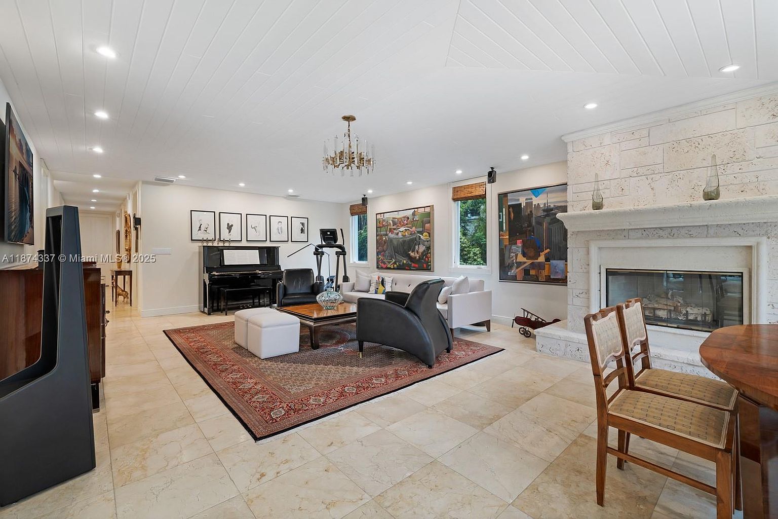 This is a spacious living room featuring a neutral color palette with a large area rug defining the seating area. The room includes a fireplace, a piano, and several pieces of art, creating a sophisticated and comfortable atmosphere. Natural light streams in through the windows, enhancing the room's inviting ambiance.