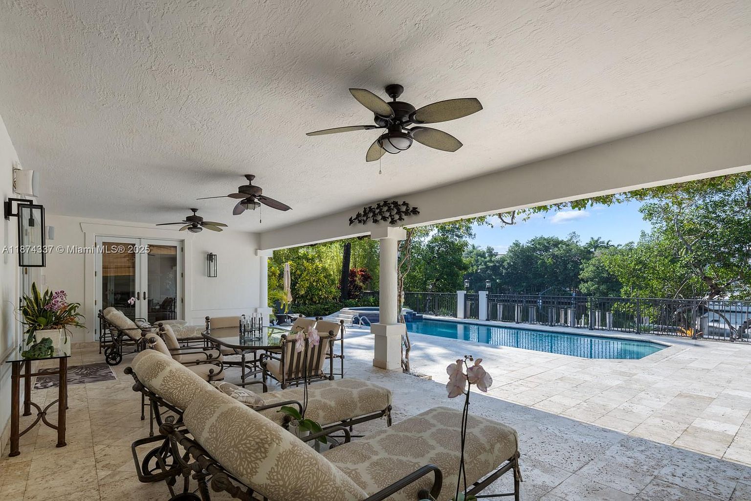 This is a view of a covered patio area with lounge chairs and a dining table, overlooking a swimming pool. The patio features travertine flooring, ceiling fans, and decorative elements. The pool is surrounded by a black metal fence and lush greenery, creating a relaxing outdoor space.