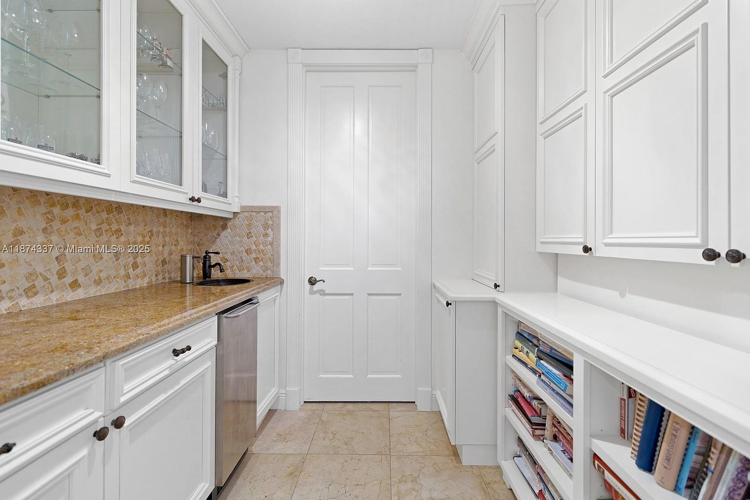 This is an interior shot of a well-organized pantry. The pantry features white cabinetry with dark hardware, a countertop with a sink, and open shelving filled with books. The floor is tiled, and a white door is centered in the background, creating a clean and functional space.