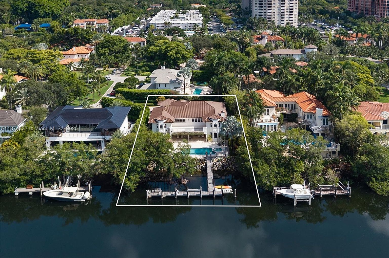 This aerial shot showcases a luxurious waterfront property with a large house, a private dock, and a swimming pool. The house features a brown tile roof and a light-colored exterior, surrounded by lush greenery and other upscale homes in the neighborhood. The image emphasizes the property's prime location and high-end amenities.