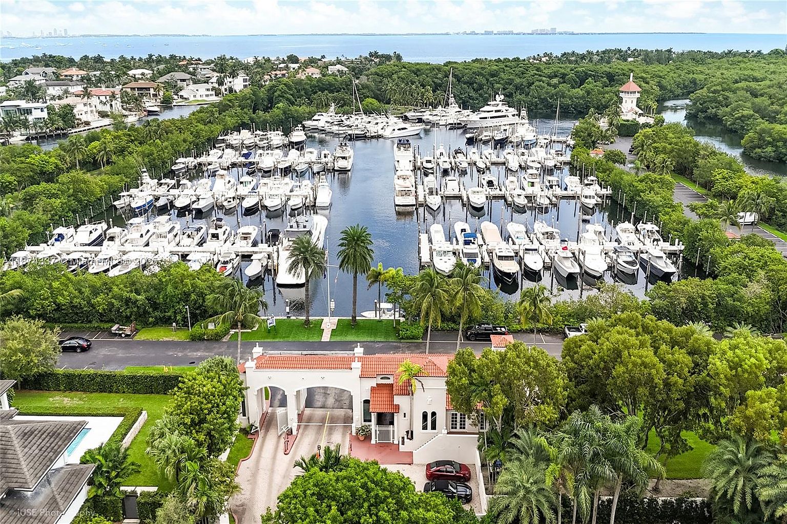 This aerial shot showcases a luxurious waterfront property with a private marina filled with numerous boats. The property features a grand entrance with a red-tiled roof and lush landscaping, creating an exclusive and serene atmosphere. The view extends to the ocean in the background, emphasizing the prime location and upscale lifestyle.