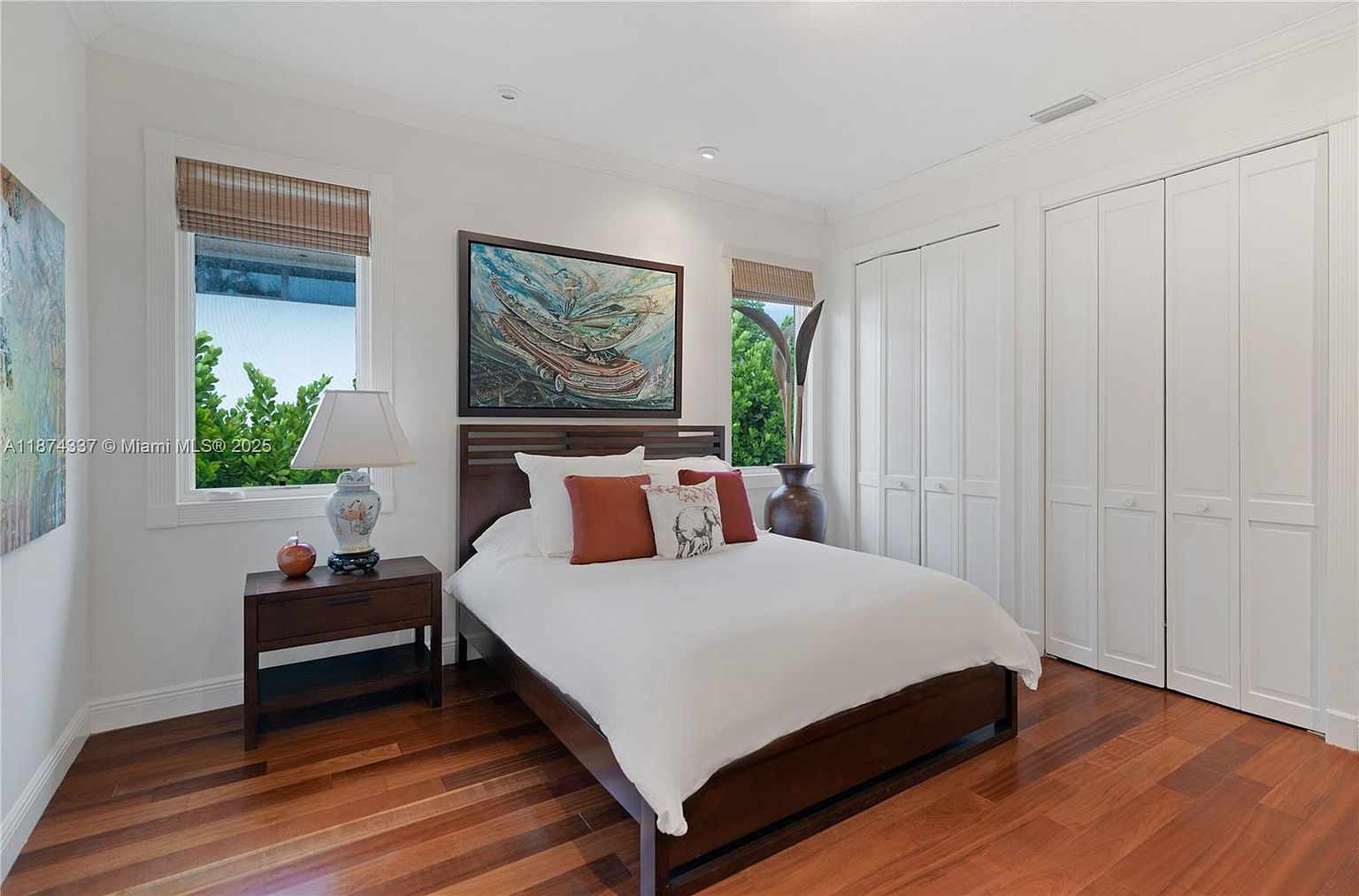 This is a well-lit bedroom featuring a dark wood bed frame with white linens and decorative pillows. A framed painting hangs above the bed, and a wooden nightstand with a lamp sits to the left. The room has hardwood floors and white closets, creating a clean and inviting space.