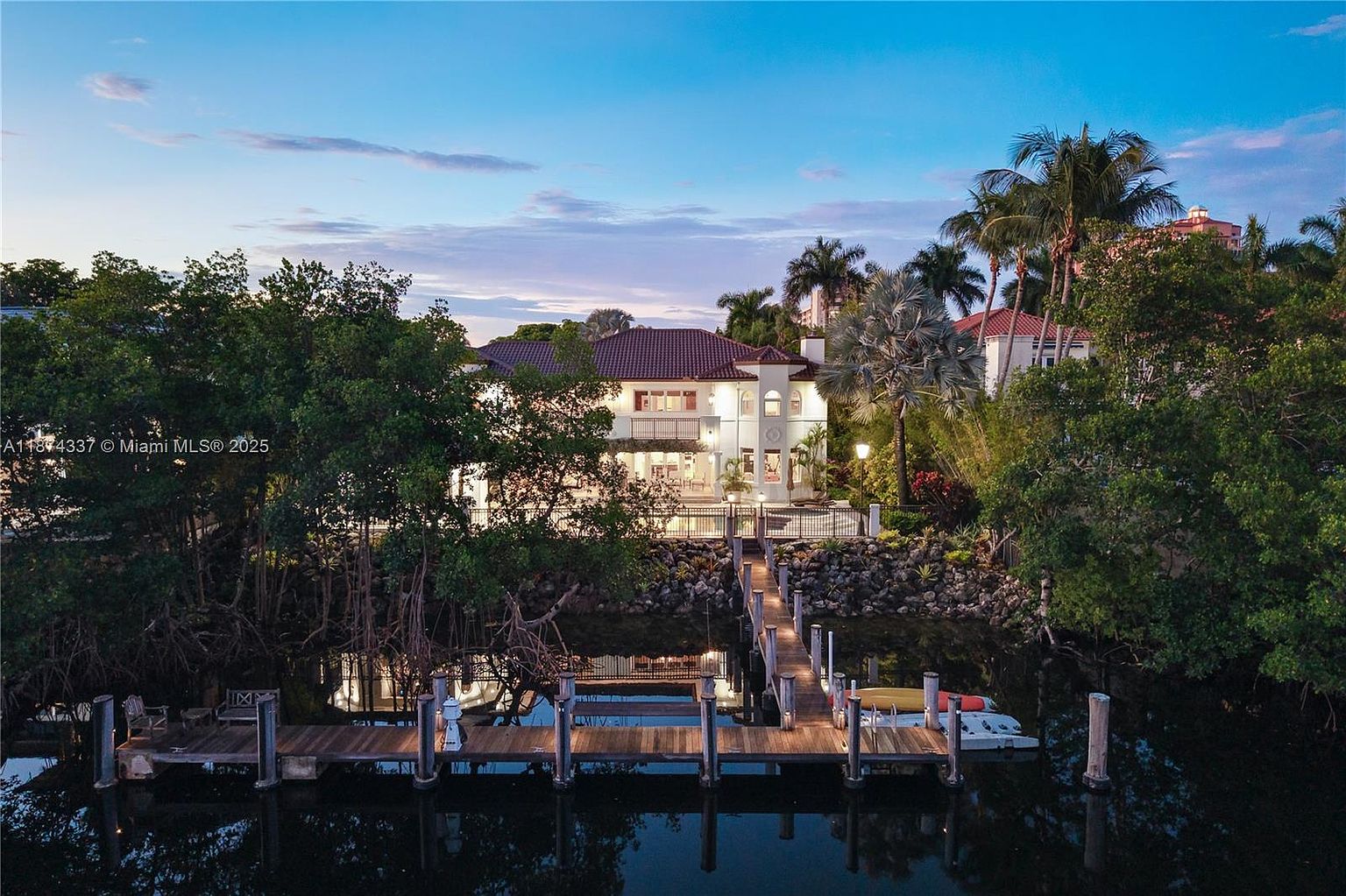 This image showcases the rear exterior of a luxurious waterfront home at dusk. The property features a multi-level design with a red tile roof, white facade, and a private dock extending into the water. Lush landscaping surrounds the home, enhancing its privacy and appeal, while the dock provides direct access to the water for boating and other recreational activities.