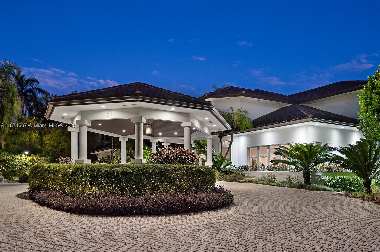 This image showcases the grand entryway of a luxurious home, featuring a covered portico with white columns and a dark tiled roof. The driveway is paved with brick, leading to a meticulously manicured circular garden with lush greenery and colorful plants. The house itself is painted white, with well-lit windows and a dark roof, exuding an elegant and welcoming ambiance.