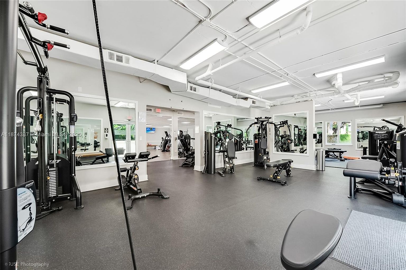 This is an interior shot of a well-equipped home gym. The space features various weight machines, exercise bikes, and benches, all set on a dark rubber floor. Mirrors line the walls, enhancing the sense of space and allowing for form checks during workouts. The room is brightly lit with overhead fluorescent lights, creating an inviting atmosphere for fitness activities.