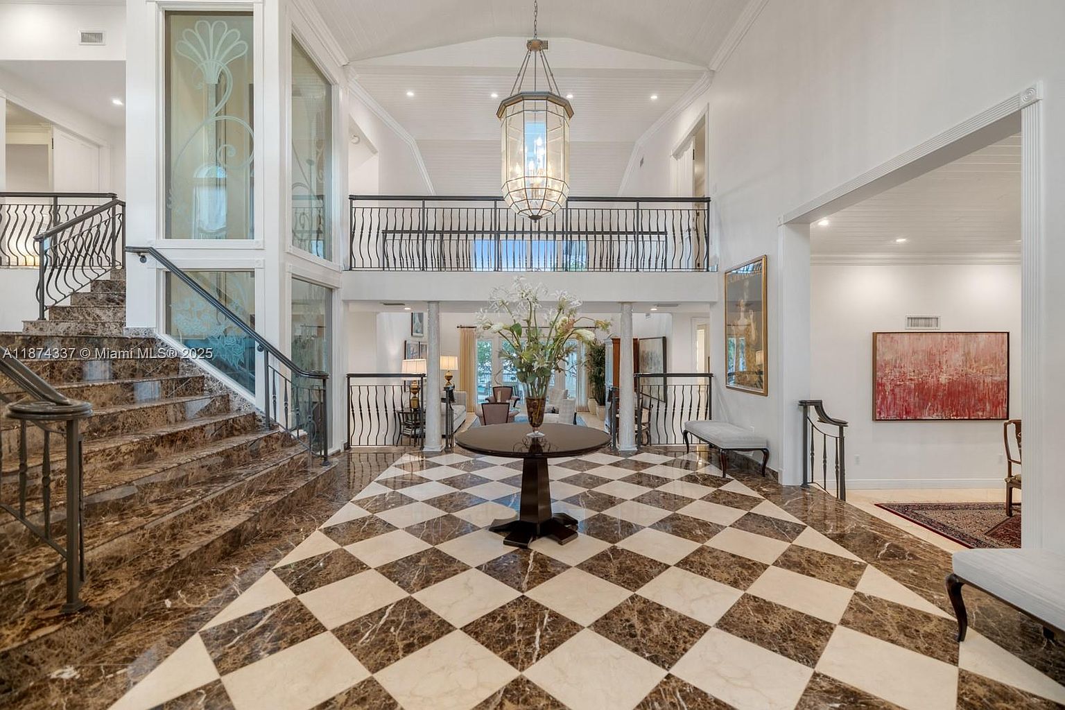 This grand foyer features a striking black and white checkered marble floor, a sweeping staircase with wrought iron railings, and a soaring two-story ceiling with a large chandelier. The space is bright and airy, with white walls and ample natural light, creating an elegant and inviting entrance. A round table with a floral arrangement serves as a focal point.