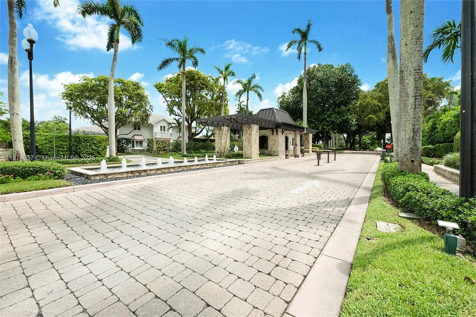 The image showcases a community entrance with a paved brick road leading to a guardhouse structure with a dark roof and stone pillars. A water fountain feature runs along the left side of the road, and palm trees line the area, creating a tropical and upscale ambiance. The scene is captured from a ground-level perspective, emphasizing the well-maintained landscaping and architectural details.