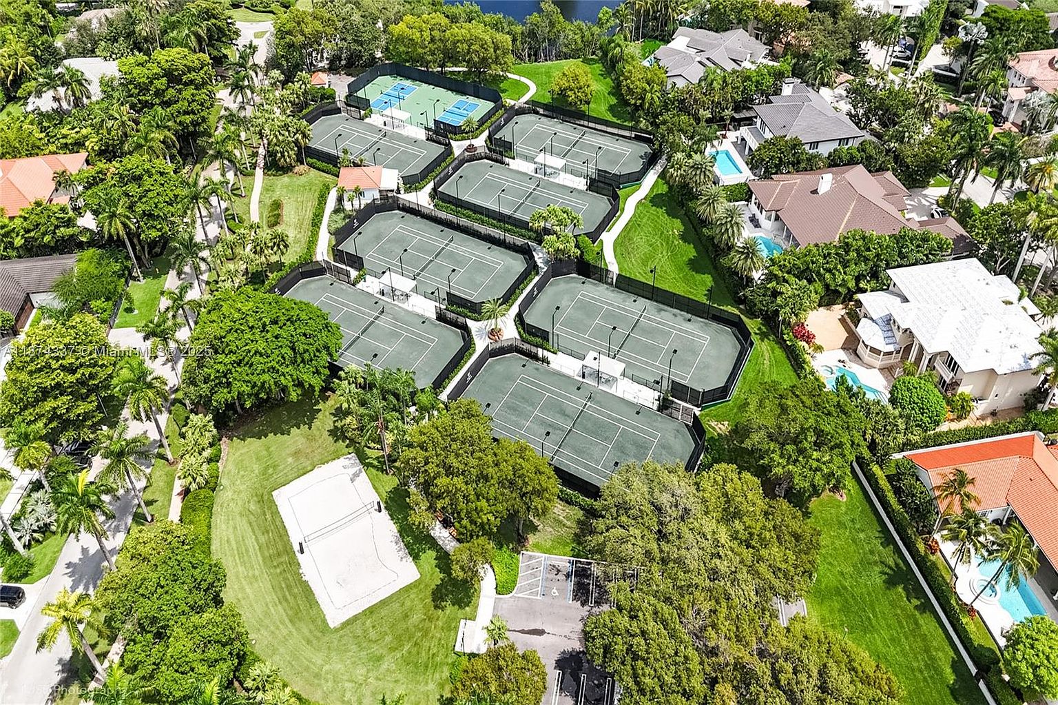 This aerial view showcases a community tennis complex with multiple well-maintained courts surrounded by lush greenery and mature trees. The courts are neatly arranged with dark fencing, and a nearby parking area and recreational space add to the amenity's appeal. The scene conveys a sense of upscale recreational opportunities within the community.