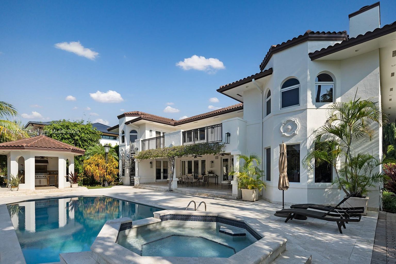 This image showcases the rear exterior of a luxurious home, emphasizing the pool and spa area. The pool is a clear blue, complemented by a tiled spa. The house features a white facade with dark brown roofing, a balcony, and lush landscaping, creating an inviting and upscale outdoor living space.