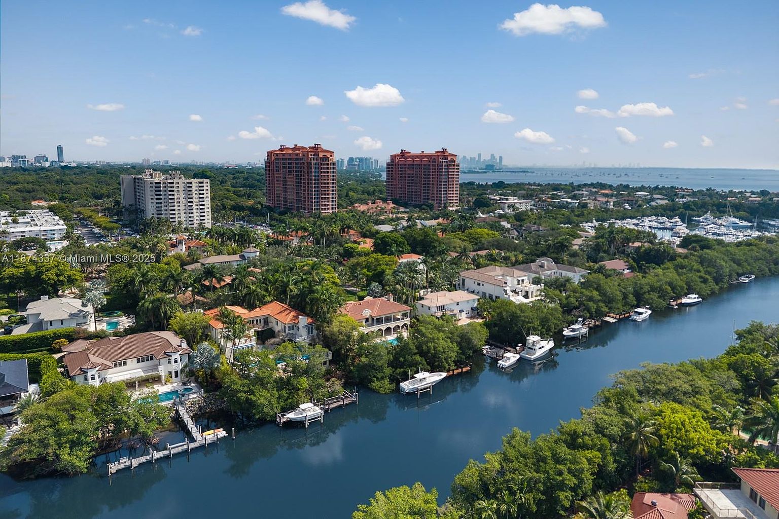 This aerial view showcases a luxurious waterfront residential area with large homes, private docks, and lush greenery. The scene includes a winding waterway, several boats, and high-rise buildings in the background, suggesting a prime location with both natural beauty and urban convenience. The overall impression is one of affluence and tranquility.