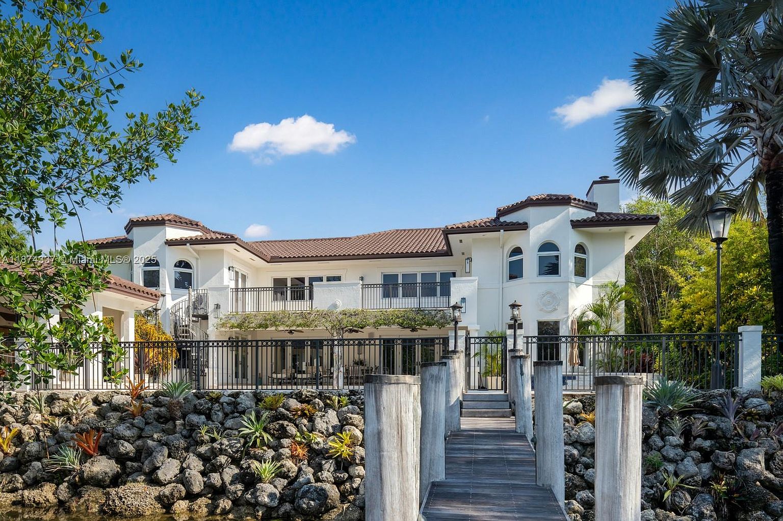 This image showcases the rear exterior of a luxurious waterfront property. The house features a white facade, a terracotta tile roof, and multiple balconies with wrought iron railings. A wooden dock extends into the water, and the landscaping includes rock features and lush greenery, creating a serene and upscale ambiance.