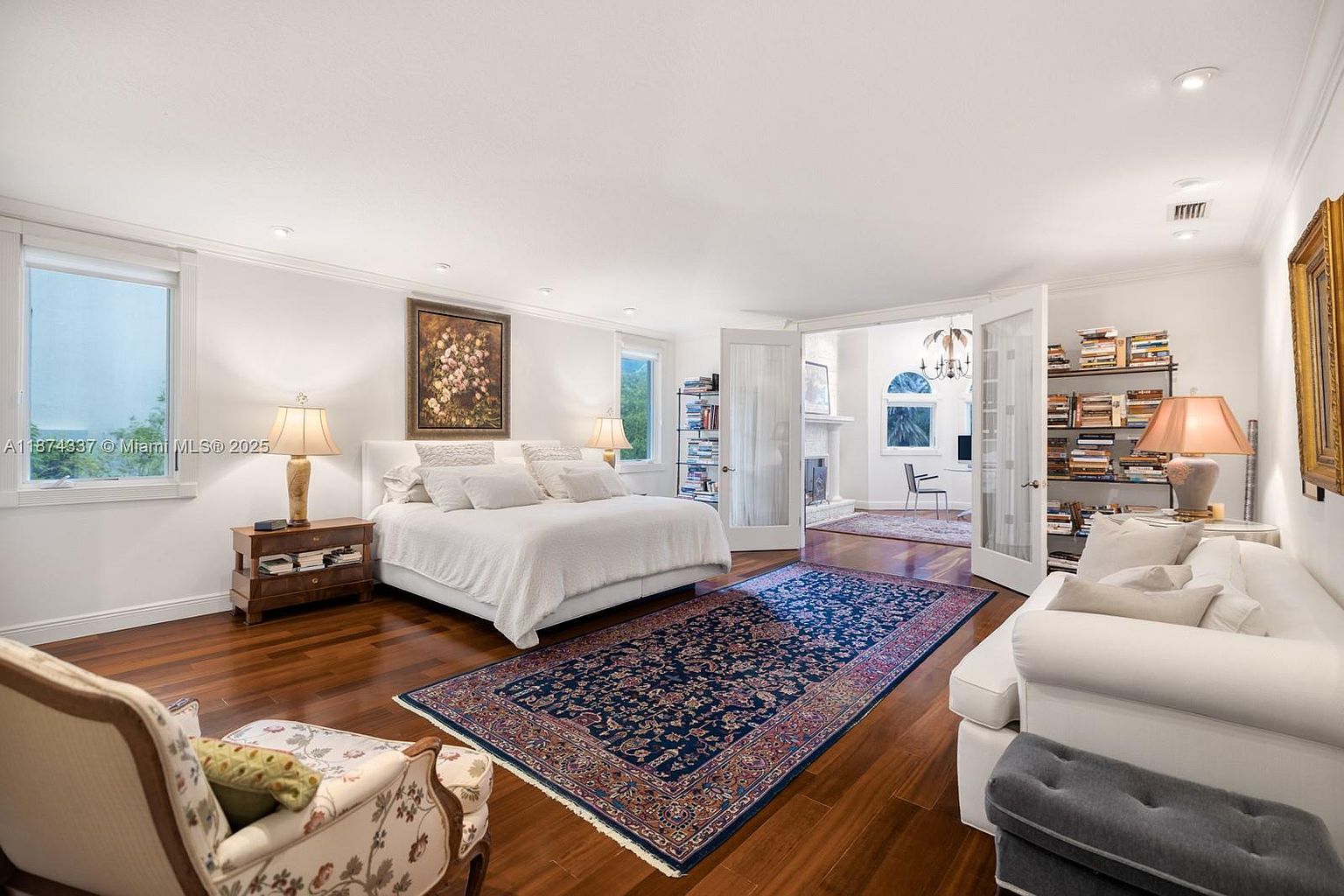 This is a spacious primary bedroom featuring hardwood floors, white walls, and a large bed with white linens. The room includes a seating area with a white sofa and a decorative armchair, as well as a large area rug. The room opens into another room through double doors, and there are windows providing natural light.