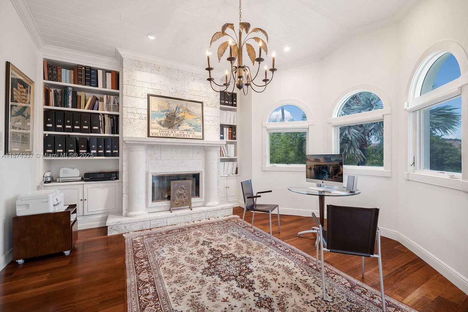 This is an interior shot of a home office or study, featuring a fireplace with a decorative mantel, built-in bookshelves, and a round glass-top desk with two chairs. The room is well-lit with natural light from arched windows, and a patterned rug covers the hardwood floor. A chandelier hangs from the ceiling, adding a touch of elegance to the space.